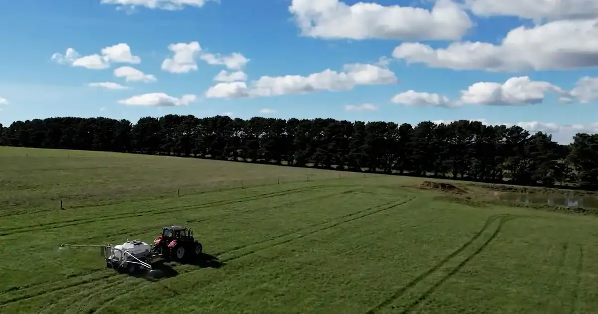 Tractor towing a Tow and Fert machine across a green pasture, spraying, with a tree line and blue sky in the background.