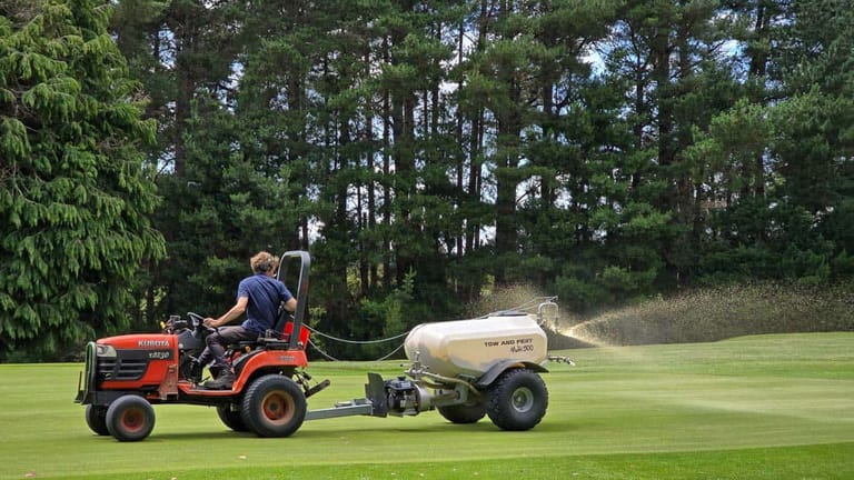 A groundskeeper driving a tractor towing a Tow and Fert Multi 500 sprayer across a golf course, spraying liquid fertiliser onto the grass with pine trees in the background.