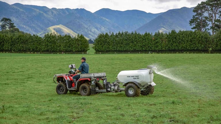 A farmer riding a quad bike towing a Tow and Fert Multi 500 sprayer across a green pasture, spraying liquid fertiliser with mountains in the background.