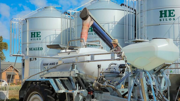 Tow and Fert MultiCrop 4000 being filled through its top filling hatch using an auger, with large fertiliser storage silos in the background.