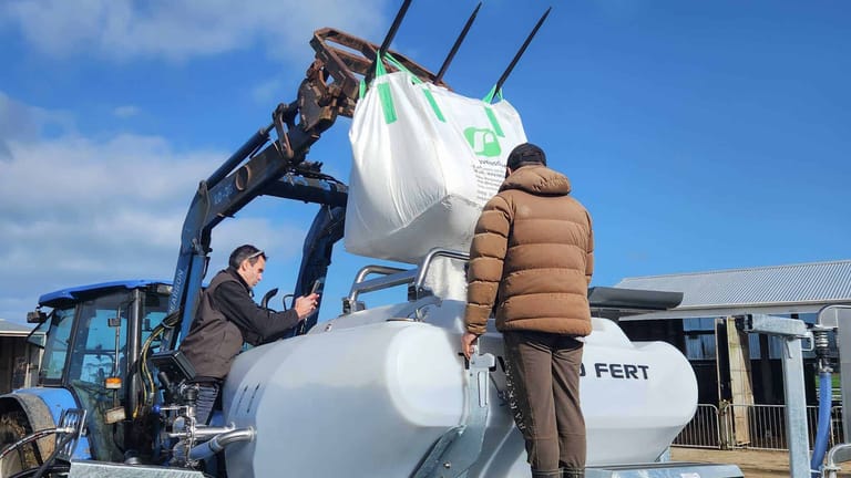 Two operators loading granular fertiliser into a Tow and Fert Multi 2800 from a bulk bag lifted by a tractor-mounted front-end loader.