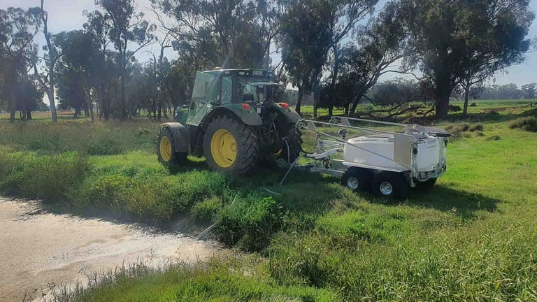 A green tractor towing a Tow and Fert sprayer, drawing water from a pond through a suction hose to fill its tank.