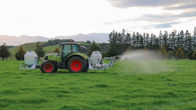 A green tractor towing a Tow and Fert Multi 1200 sprayer across a green pasture, applying liquid fertiliser through a spray boom.